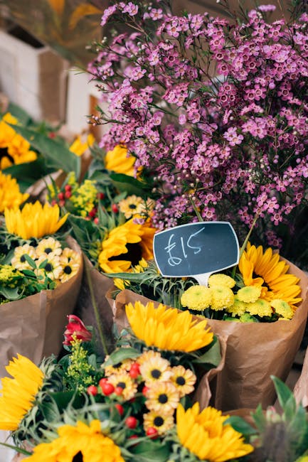 A vibrant assortment of fresh flowers displayed at a florist stall, featuring bright yellow sunflowers, small white daisies with yellow centers, and clusters of delicate pink flowers. The bouquets are
