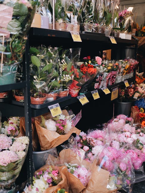 An indoor display of fresh floral arrangements at a florist shop, with various bouquets and potted plants arranged on black metal shelves. The top shelf showcases potted green plants and orchids, wrap