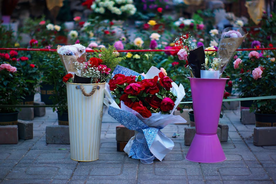 A floral display featuring a bouquet of deep red roses wrapped in white and blue paper, placed on a paved surface outdoors. To the left, a tall white and gold striped vase holds arrangements of small