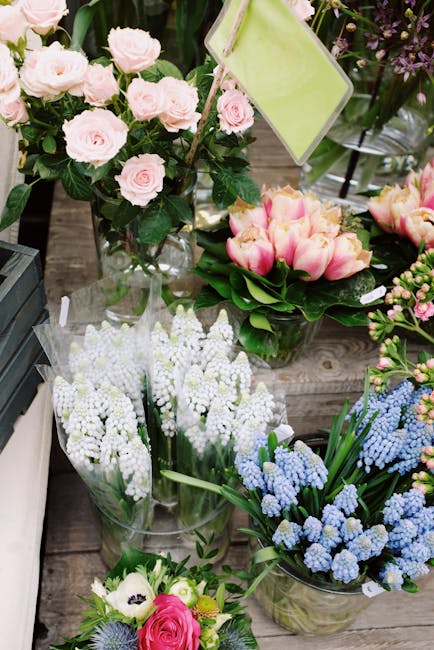 A variety of fresh floral arrangements displayed on a wooden surface, including a bouquet of pale pink roses with lush green leaves in a clear glass vase, a separate arrangement of pink and cream tuli