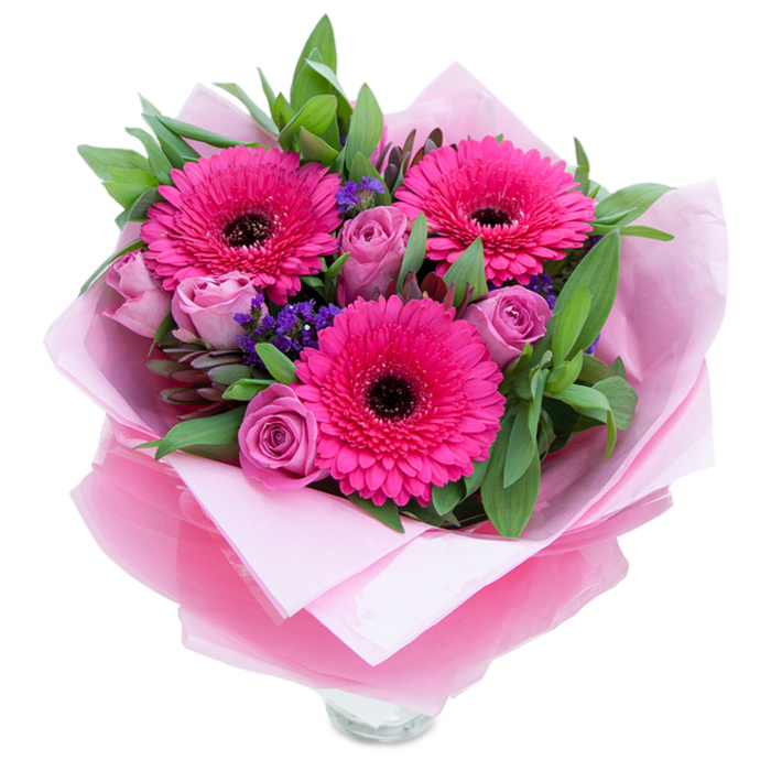 This arrangement captures a playful yet elegant spirit through three fuchsia Gerbera daisies that burst with layered petals and dark, defining centres; they act as joyful focal points amid a softer chorus of flowers. The roses here are a muted lavender-pink, their petals still forming tight, velvety spirals that suggest freshness and a tender sweetness. Scattered between the main blooms, small bunches of purple statice add a cool, papery texture and a subtle depth that keeps the eye moving through the design. Surrounding everything, broad green leaves-likely salal with hints of eucalyptus-offer a crisp, garden-fresh backdrop and a faintly aromatic lift that balances the floral notes. The bouquet is wrapped with intention: an inner sheet of pale pink, matte tissue gives a soft, protective cradle while an outer layer of translucent fuchsia-pink cellophane, slightly crinkled, provides a glinting surface that reinforces the pink theme and gives the whole piece a polished presentation. The lower section exposes the top rim of a clear glass vase, suggesting the flowers are ready to be arranged for immediate display. In the context of Shepherd's Bush-whether arriving near Westfield London, along Uxbridge Road, or at a cosy home by Goldhawk Road-this bouquet reads as a thoughtful gift for birthdays, anniversaries, or to brighten a room after a long day. It combines visual energy with gentle fragrance and expert handling, the kind of composition a local florist would make to convey affection and celebration.